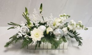 A mix of white flowers with holiday greens arranged in a mirrored rectangle container, with keepsake ornaments.
