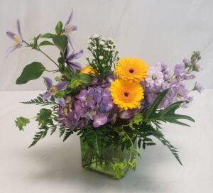 Soft lavender colored flowers and yellow germinis arranged in a square green glass container