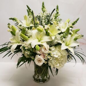 An all white flower arrangement with beautiful lilies, snapdragons, and hydrangeas in a clear vase.