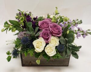 Low textured arrangement in a wooden planter box, features roses, stock, thistle, and succulents.