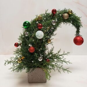 A curved whimsical holiday tree with holiday ornaments in a square container