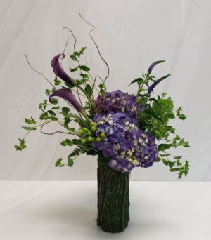 An organic mix of green and purples with curly willow arranged in a moss-wrapped vase