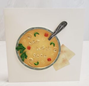 A 6"x6" quilling card that says, "Get Well Soon." Features a quilled bowl of soup and a spoon.