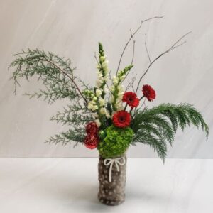 A holiday arrangement of red, white, and green blooms with holiday greens and birch branches in a cylinder bundled in a wine gift bag